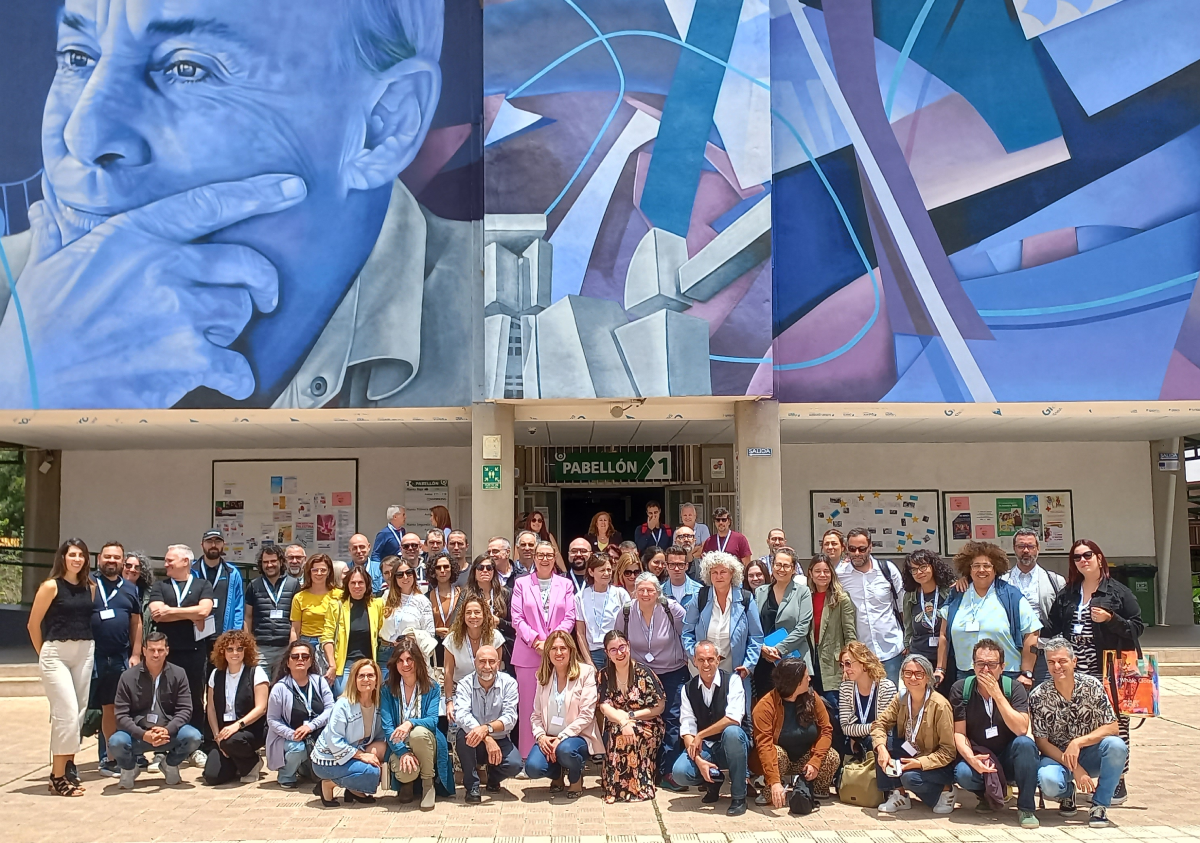 Grupo numeroso de personas posando para una fotografía, con un gran mural sobres sus cabezas que incluye el rostro de una persona, con una mano sobre el mentón y gesto pensativo, a su izquierda